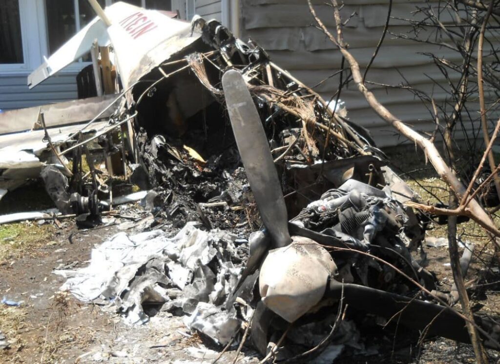 Charred aircraft engine and propeller remain embedded near the side of a house. The fuselage is severely burned, with the cabin structure almost completely destroyed.